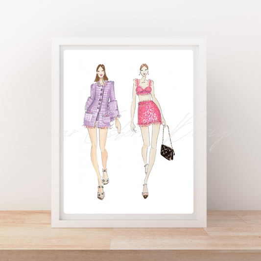 A framed fashion illustration print leaning against a wall on a light wood surface. The artwork shows two stylized women in pink and purple outfits, and the print is enclosed in a white frame.